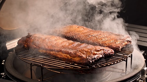 Ribs on the grill with billowing smoke coming from the SloRoller smoking accessory below