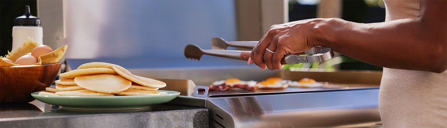 Bacon, eggs, and sausages grilling on a large flat top gas grill
