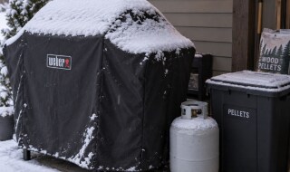Snow-dusted covered Weber grill next to a spare propane tank and a sealed pellet bin
