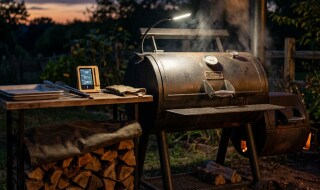 Offset smoker running at dusk with a grill light on the cook chamber and a covered woodpile on a rack nearby