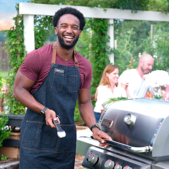 Smiling man in BBQ apron standing outdoors with friends gathered around a table in a lush, green garden setting.